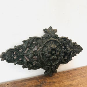 Op de afbeelding: Een sierlijk, donkergroen architecturaal element van gietijzer met een centraal bloemenmedaillon en krullende bladmotieven. Het decoratieve stuk is gemonteerd op een houten oppervlak. Het is waarschijnlijk een decoratief accent.