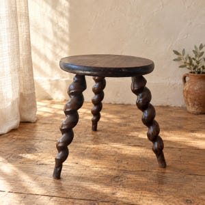 May include: A dark brown wooden stool featuring a smooth, circular top and three distinct legs carved into a spiral, twisted shape. The stool has a polished finish that highlights the natural grain and deep color of the wood throughout the structure.