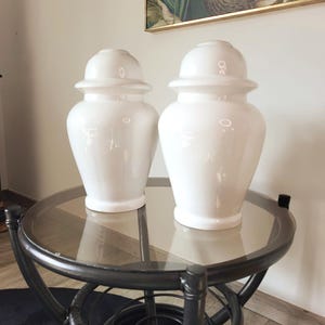 May include: Two white ceramic decorative jars with lids sit on a round glass-topped table with a black metal frame. The jars have a glossy finish and a classic urn shape. A framed artwork hangs in the background.