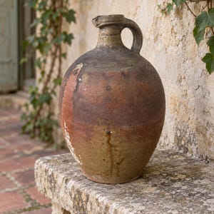 Peut inclure: Une cruche en céramique rustique et bombée avec une anse unique et un col étroit. L'extérieur présente une finition patinée et multicolore dans des tons de brun terreux, de vert olive sourd et de gris anthracite, avec des marques d'usure.