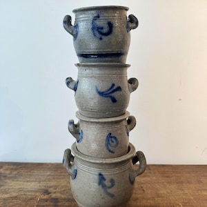 Puede incluir: Una pila vertical de cuatro macetas de gres gris, cada una con dos asas laterales y marcas decorativas pintadas a mano en azul. Las macetas tienen un acabado texturizado y moteado, con una forma cilíndrica y sutiles crestas horizontales.