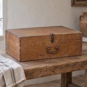 May include: A rectangular wooden storage box with a weathered brown finish, featuring visible finger joints at the corners, a central metal latch on the front lid, and a curved metal handle attached to the front panel for carrying purposes.