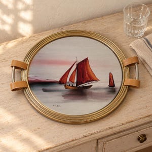 Peut inclure: Un plateau de service rond avec un cadre doré et des poignées en cuir beige. Le centre présente une illustration peinte de deux voiliers aux voiles rouge foncé sur une mer calme et claire sous un ciel rose et gris doux, signé par l'artiste.