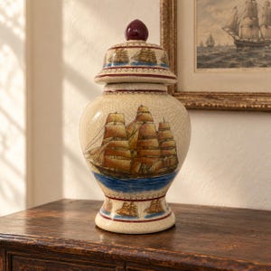Può includere: Un vaso in ceramica con finitura craquelé, decorato con un veliero dipinto a mano che naviga su acqua blu. Il vaso include un coperchio abbinato con un pomello rosso scuro e bordi nautici decorativi con piccoli motivi di navi alla base.