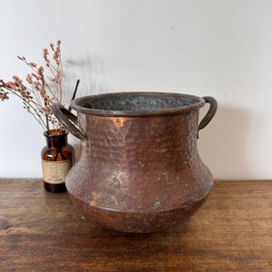 Könnte beinhalten: Ein gehammerter Kupfertopf mit zwei kleinen Griffen. Der Topf hat einen breiten, abgerundeten Körper und einen leicht ausgestellten Rand. Das Kupfer hat eine reiche, gealterte Patina mit grünen Oxidationsbereichen. Der Topf steht auf einer Holzoberfläche.