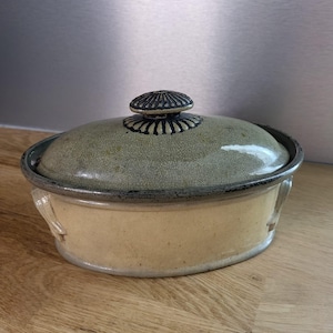 Peut inclure: Un plat à casserole en céramique ovale avec un couvercle. Le plat a une base beige clair et un couvercle vert-gris. Le couvercle est orné d'une poignée décorative. Le plat est posé sur une surface en bois.