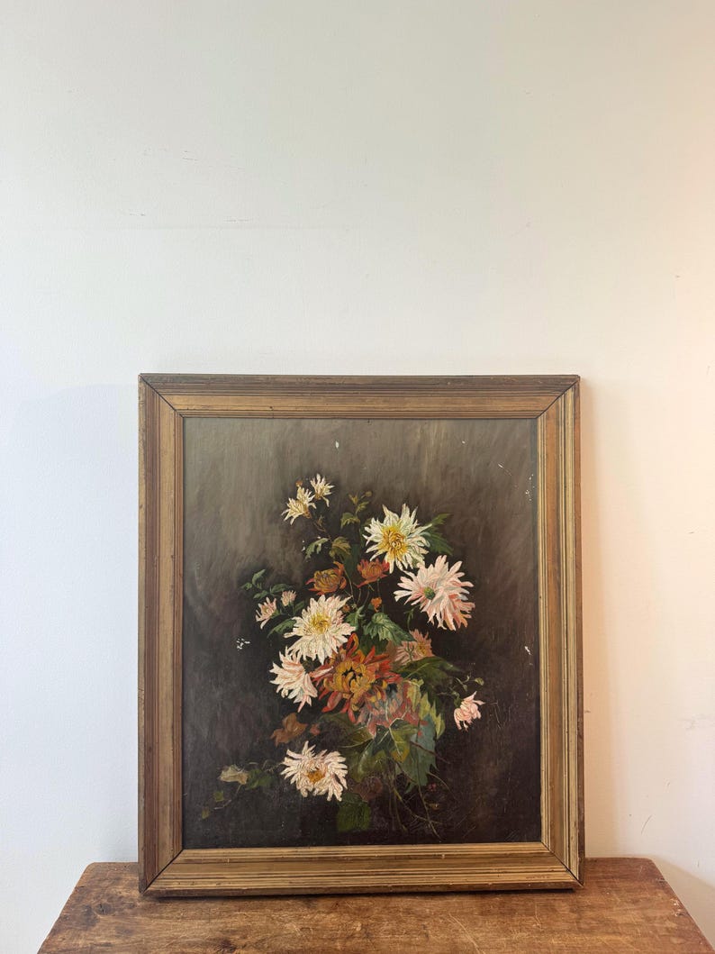 Op de afbeelding: Een antiek, ingelijst bloemenschilderij. Het kunstwerk toont een boeket witte, gele en oranje bloemen tegen een donkere achtergrond. De houten lijst heeft een warme, gouden tint, wat de vintage uitstraling versterkt.