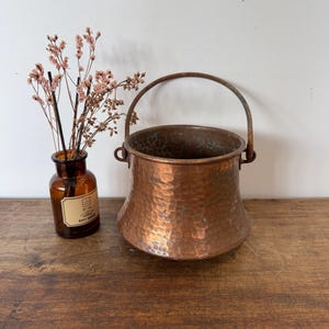Puede incluir: Un caldero de cobre martillado con asa, sobre una superficie de madera. El caldero tiene una base ancha y una parte superior estrecha. A la izquierda del caldero hay una pequeña botella de vidrio marrón con flores secas.