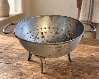 May include: A weathered, round metal colander featuring a silver-toned finish with visible signs of oxidation and wear. The bowl includes two side handles, a series of drainage holes throughout the basin, and stands on three short, sturdy metal legs.