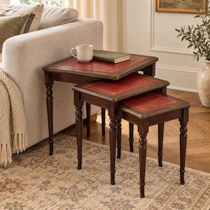 Op de afbeelding: Een set van drie bijzettafels van donkerbruin hout met rode lederen inlays op de rechthoekige bladen. Elke tafel is voorzien van vier gedraaide poten en goudkleurige decoratieve randdetails rondom de lederen oppervlakken.