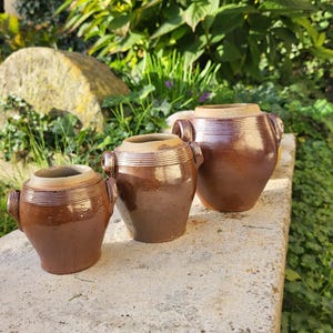 May include: Three brown ceramic pots of different sizes are displayed. Each pot has a rounded body, a slightly flared rim, and two small handles. The pots have a glossy finish and are on a stone surface, with greenery in the background.