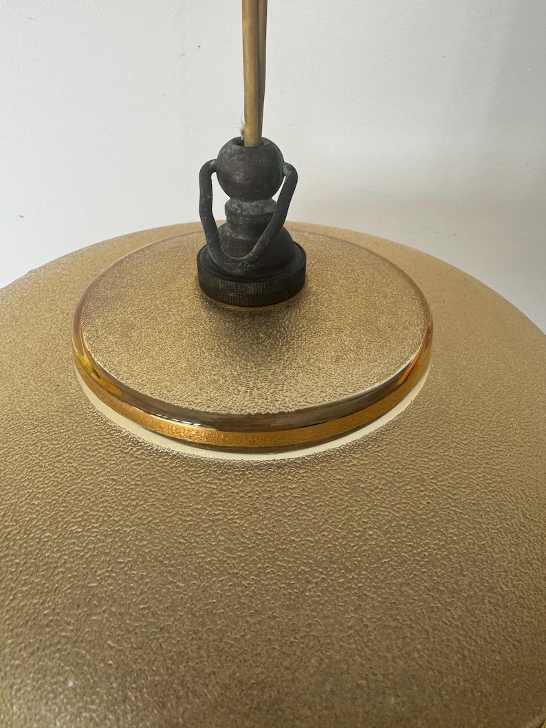 May include: A gold-colored glass pendant light fixture with a textured surface and a gold trim. The fixture has a simple, round shape and a metal hook for hanging.