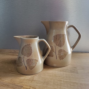Peut inclure: Deux pichets en céramique avec un motif de feuilles. Le plus grand pichet est brun avec un motif de feuilles brun clair. Le plus petit pichet est brun clair avec un motif de feuilles brun foncé.