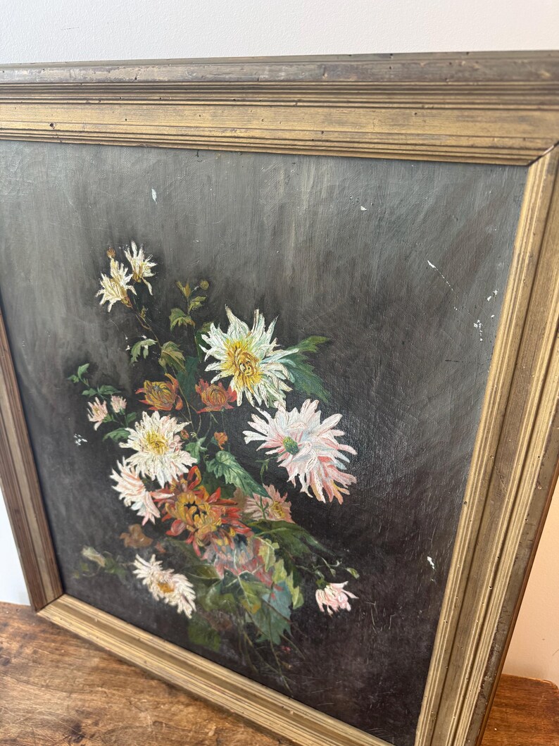 Op de afbeelding: Olieverfschilderij van een boeket bloemen in een goudkleurige houten lijst. De bloemen zijn wit, roze en oranje, met groene bladeren tegen een donkergrijze achtergrond. De lijst heeft een getextureerd oppervlak.