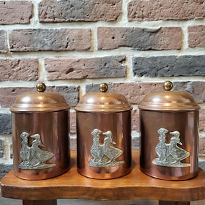 Puede incluir: Conjunto de tres botes de cobre con diseños de gansos plateados. Los botes están dispuestos en una fila en un estante de madera.