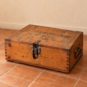 Könnte beinhalten: Eine rechteckige Aufbewahrungsbox aus Holz mit sichtbaren Fingerzinken an den Ecken, einem Metallverschluss an der Vorderseite und Metallgriffen an den Seiten. Das Holz hat ein natürliches braunes Finish mit verblasstem schwarzem Text auf dem Deckel.
