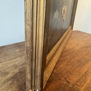 Op de afbeelding: Een vintage schilderij in een goudkleurige houten lijst. De lijst heeft een gedetailleerd, gefluteerd ontwerp. Het kunstwerk heeft gedempte kleuren en een zichtbare textuur. Het schilderij wordt getoond op een donker houten oppervlak.