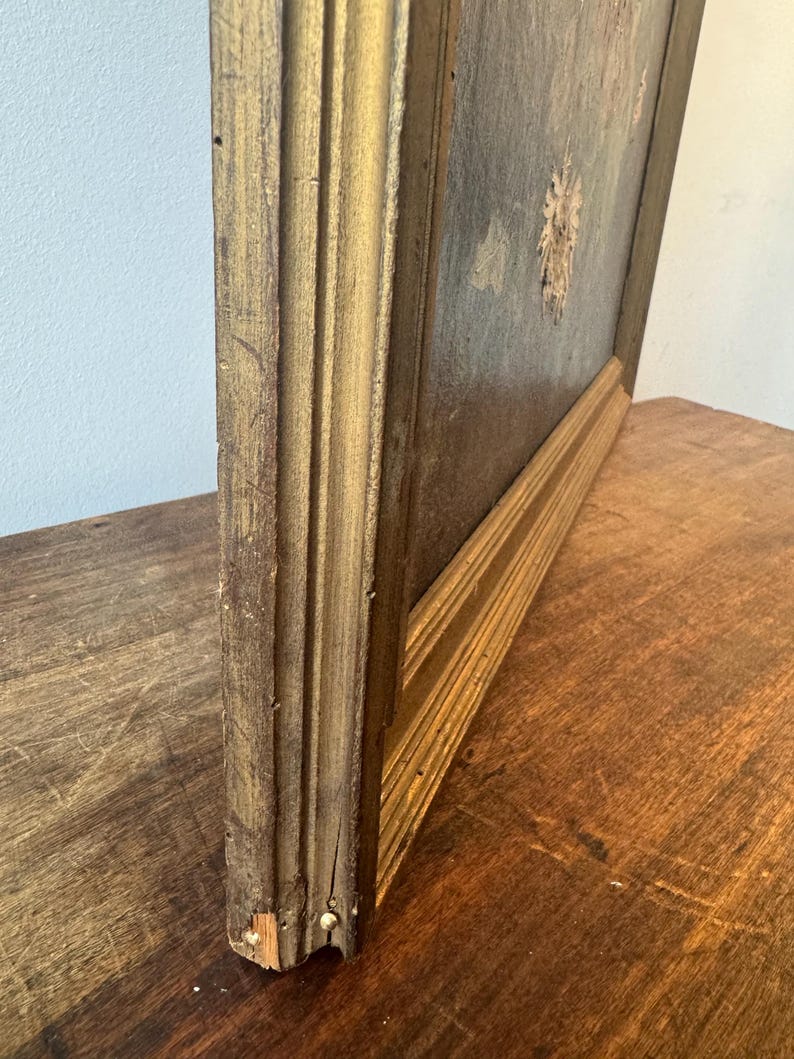 Op de afbeelding: Een vintage schilderij in een goudkleurige houten lijst. De lijst heeft een gedetailleerd, gefluteerd ontwerp. Het kunstwerk heeft gedempte kleuren en een zichtbare textuur. Het schilderij wordt getoond op een donker houten oppervlak.