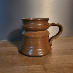 Peut inclure: Une tasse en céramique brune avec une base large et un bord évasé. La tasse a une surface texturée et une anse.
