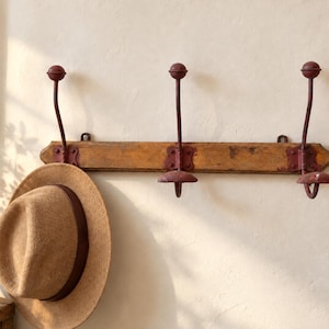 May include: A wall-mounted coat rack featuring a horizontal wooden plank with three dark red metal hooks, each topped with a spherical finial. A tan felt hat with a dark brown band hangs from the leftmost hook on the rack.