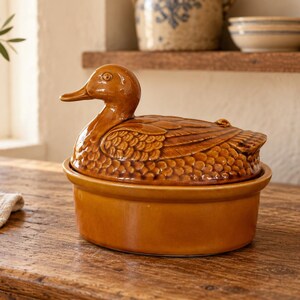 Op de afbeelding: Een glanzende, amberkleurige keramische ovenschaal met een deksel in de vorm van een eend. Het deksel is voorzien van een gevormde, driedimensionale eendenfiguur met gedetailleerde verenstructuren, rustend op een gladde, ovale basis met glazuur.