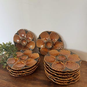 May include: A collection of round ceramic oyster plates featuring a glossy brown glaze and a molded shell pattern with six individual compartments surrounding a central circular well. The plates are stacked in two separate piles on a surface.