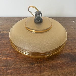 May include: A round, amber-colored glass ceiling light fixture with a gold trim. The light fixture has a textured surface and a simple metal hook.