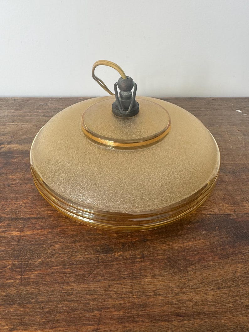 May include: A round, amber-colored glass ceiling light fixture with a gold trim. The light fixture has a textured surface and a simple metal hook.