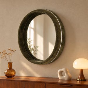 May include: A circular wall mirror featuring a thick, dark olive green velvet-textured frame. The mirror has a reflective glass surface that shows a blurred view of a window and greenery. The frame has a smooth, rounded profile.