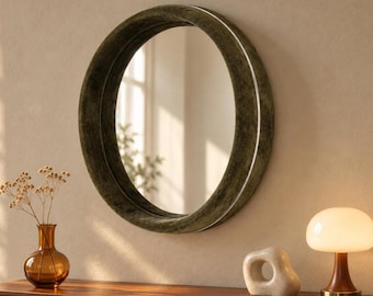 May include: A circular wall mirror featuring a thick, dark olive green velvet-textured frame. The mirror has a reflective glass surface that shows a blurred view of a window and greenery. The frame has a smooth, rounded profile.