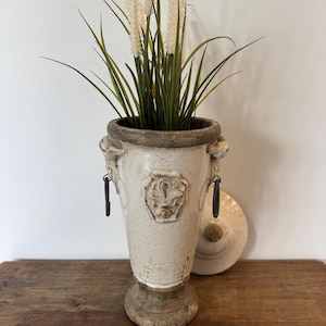 May include: A cream-coloured, antique-style urn with a distressed finish, featuring a fleur-de-lis emblem and decorative handles. The urn is filled with green and beige ornamental grass. A matching lid rests beside it.