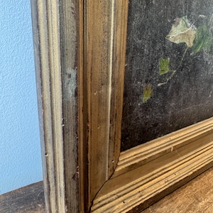Op de afbeelding: Close-up van een vintage schilderij in een goudkleurige houten lijst. De lijst heeft een verweerde afwerking. Het schilderij toont donkere kleuren met hints van groen en bruin blad. Het kunstwerk wordt getoond op een houten oppervlak.