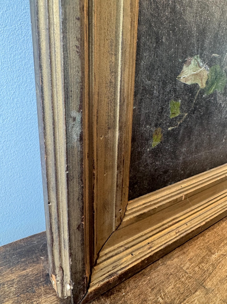 Op de afbeelding: Close-up van een vintage schilderij in een goudkleurige houten lijst. De lijst heeft een verweerde afwerking. Het schilderij toont donkere kleuren met hints van groen en bruin blad. Het kunstwerk wordt getoond op een houten oppervlak.