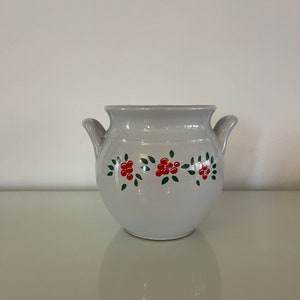 Op de afbeelding: Een witte keramische pot met twee handvatten en een bloemenontwerp met rode bessen en groene bladeren.