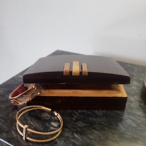 Op de afbeelding: Een donkerhouten juwelendoos met een gouden rand, open om een ​​houten interieur te onthullen. Twee gouden armbanden liggen op het oppervlak van een donkergrijs aanrecht.