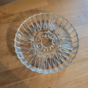 Puede incluir: Un plato de servir de vidrio transparente con un patrón de estrella. El plato tiene una sección central elevada y un borde festoneado.