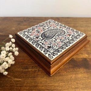 Peut inclure: Boîte carrée en bois avec un dessus en carreau de céramique décoratif. Le carreau présente un motif cachemire au centre, entouré de fleurs roses et de motifs floraux noirs et blancs. La boîte a un cadre en bois foncé.