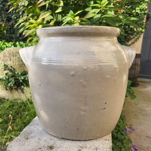 Op de afbeelding: Een grote, gebroken witte steengoed pot met twee zijhandvatten. De keramische pot heeft een brede opening en een licht getextureerd oppervlak. De pot staat op een stenen oppervlak, met groen op de achtergrond.