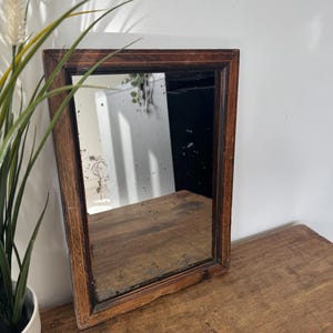 Peut inclure: Un miroir rectangulaire ancien avec un cadre en bois brun foncé. La surface du miroir présente des signes de vieillissement avec des taches et des imperfections. Il repose sur une surface en bois, avec une plante dans un pot blanc à gauche.