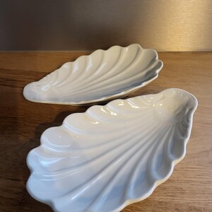Peut inclure: Deux plats de service en céramique blanche en forme de coquille. Les plats sont ovales et ont une surface texturée et nervurée qui ressemble à une coquille.