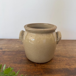 May include: A tan-colored, ceramic pot with two small handles on the sides. The pot has a rounded body and a slightly flared rim. The surface has a speckled appearance. The pot is likely used for storage or decoration.