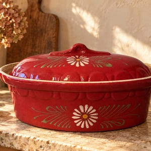 Peut inclure: Un plat à four ovale en céramique rouge profond avec un couvercle assorti. L'extérieur présente un motif de marguerite peint à la main en blanc et jaune avec des feuilles vertes. Le bord du plat et le couvercle sont festonnés, l'intérieur est beige.