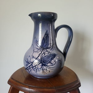 Peut inclure: Un pichet en céramique bleu et blanc avec un motif de chardon. Le pichet a une large ouverture et une anse. Il est posé sur un support en bois.