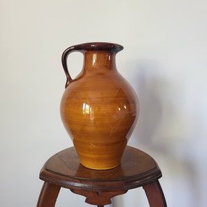 Pode incluir: Um jarro grande de cerâmica marrom com uma alça, colocado sobre uma pequena mesa de madeira escura. O jarro tem um acabamento brilhante e uma borda marrom mais escura. A mesa tem um tampo redondo e pernas curvas.