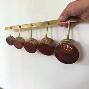 May include: Five copper saucepans with brass handles suspended from a brass bar. The pans vary in size and have a hammered texture. The copper has a reddish-brown colour, and the handles are a shiny gold colour. A kitchenware set.