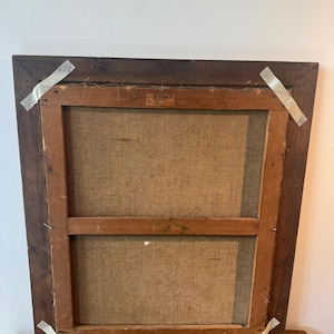 Op de afbeelding: Achterkant van een ingelijst schilderij. De donkerbruine houten lijst is rechthoekig. Het canvas is lichtbeige van kleur, gespannen over een houten frame. Het canvas is met nietjes en tape aan het frame bevestigd.