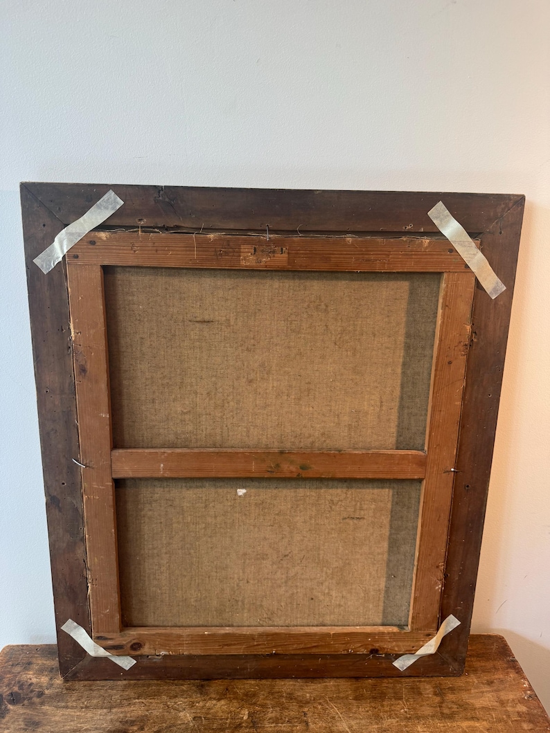 Op de afbeelding: Achterkant van een ingelijst schilderij. De donkerbruine houten lijst is rechthoekig. Het canvas is lichtbeige van kleur, gespannen over een houten frame. Het canvas is met nietjes en tape aan het frame bevestigd.