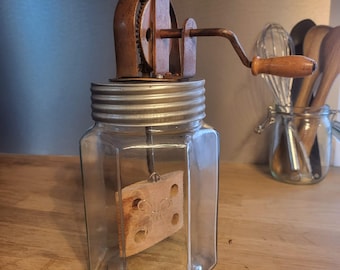 Puede incluir: Un barril de mantequilla de vidrio vintage con una paleta de madera en su interior. El barril tiene una tapa de metal y una manivela manual en la parte superior. La manivela es de metal y madera.