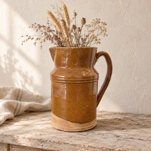 Pode incluir: Um jarro de cerâmica alto e cilíndrico com um esmalte brilhante de cor âmbar profundo e uma única asa curva. A base mostra uma secção de argila bege não vidrada. O jarro está cheio com um bouquet de caules de trigo e flores secas.