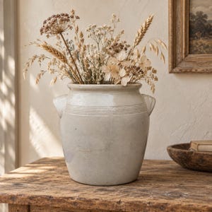 Op de afbeelding: Een lichtgrijze keramische pot met twee kleine handvatten aan de zijkanten en horizontale ribbels bij de rand, gevuld met een verscheidenheid aan gedroogde beige wilde bloemen, tarwearen en grassen voor een neutraal en textuurrijk boeket.